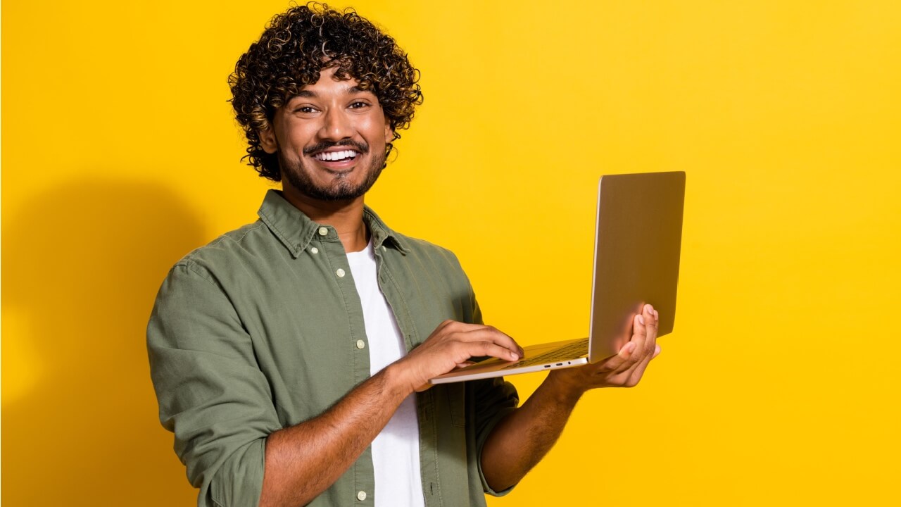 Retrato fotográfico de um homem de cabelos cacheados segurando um computador, isolado em um fundo amarelo.