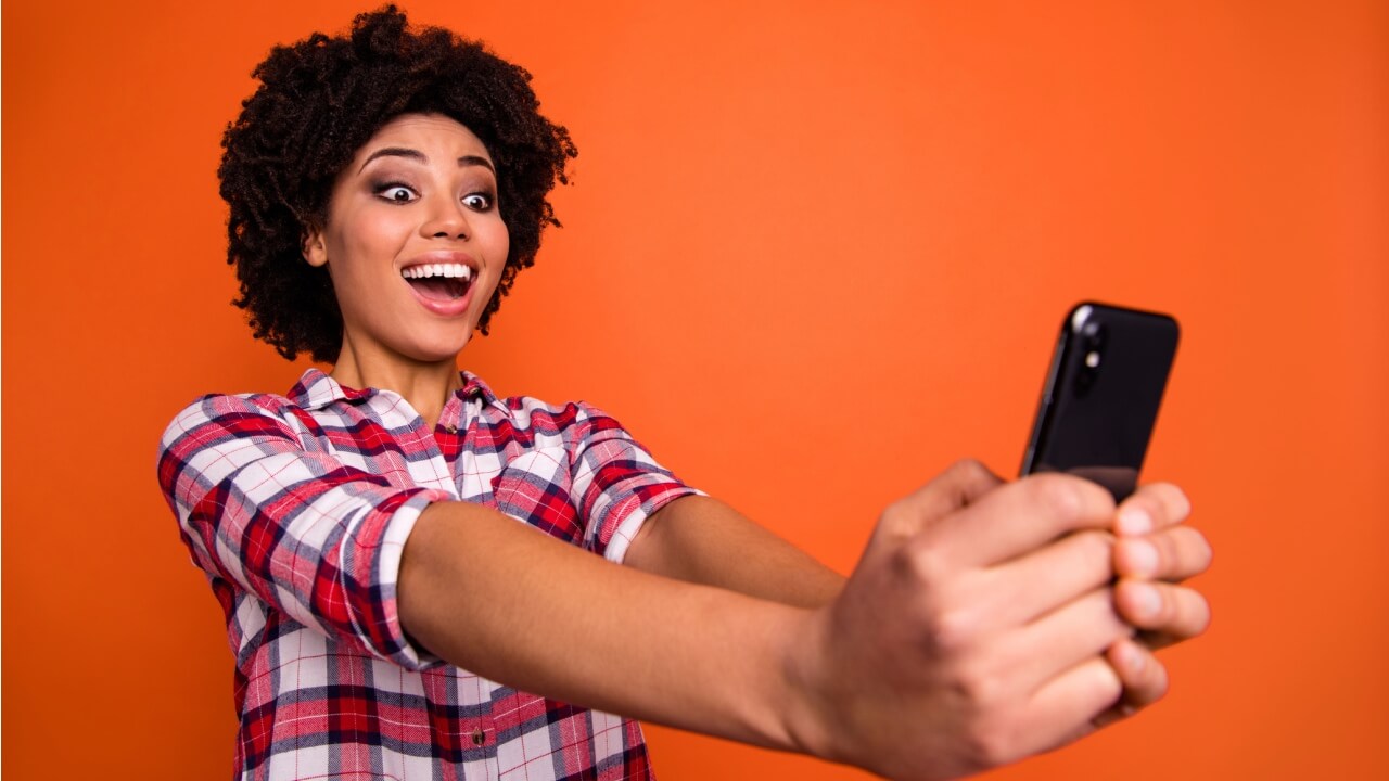 Foto de uma mulher segurando um celular, feliz, usando uma camisa xadrez casual, isolada em um fundo laranja.