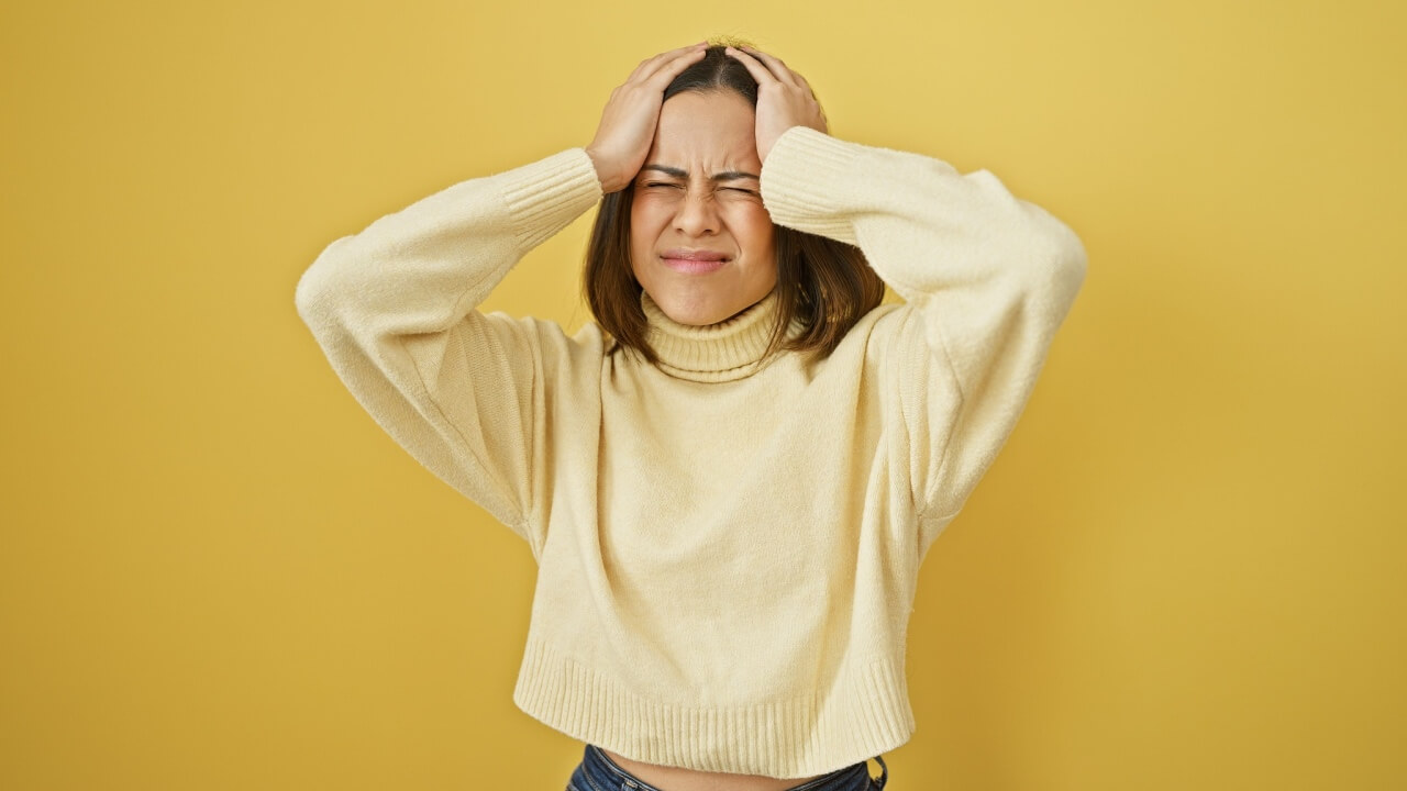 Mulher estressada com as mãos na cabeça sobre um fundo amarelo simples.