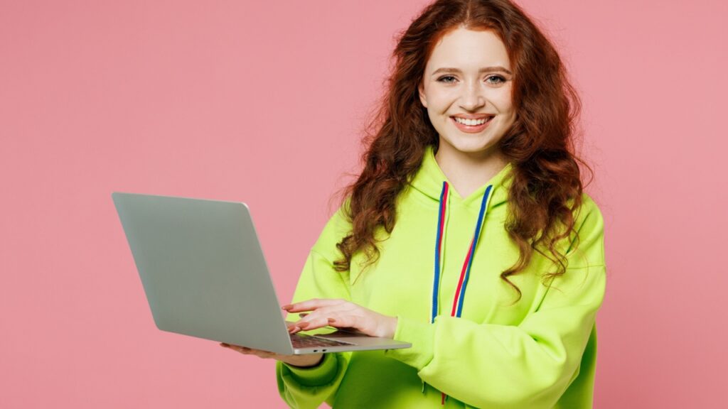 mulher de cabelo vermelho com um corta vento verde limão carregando um notebook nas mãos em fundo rosa