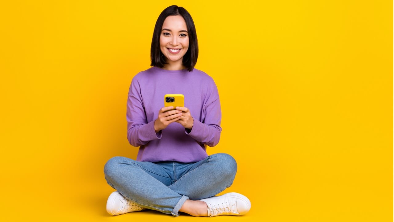 Foto de corpo inteiro de uma mulher otimista segurando um celular, isolada em um fundo amarelo.