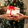 imagem de duas mãos tocando uma caixa de presente de natal com uma árvore de natal desfocada ao fundo