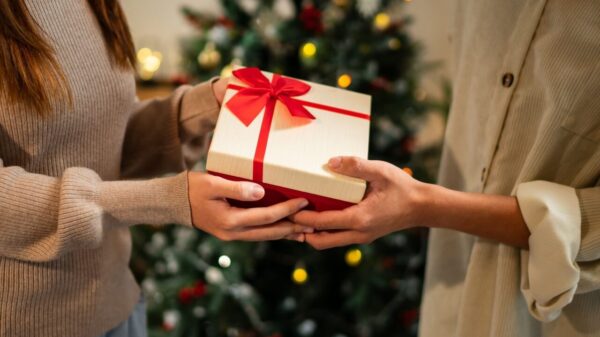 imagem de duas mãos tocando uma caixa de presente de natal com uma árvore de natal desfocada ao fundo