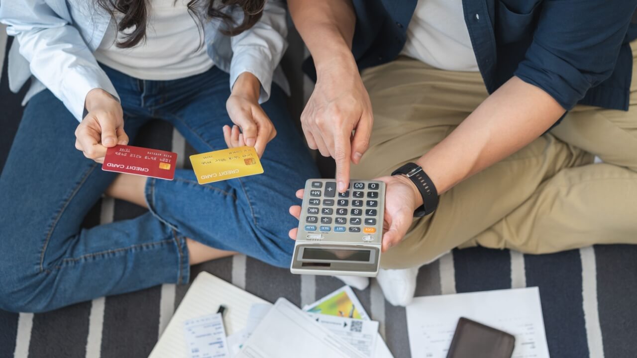 casal sentado no chão fazendo cálculos de dívidas com calculadora e cartão na mão