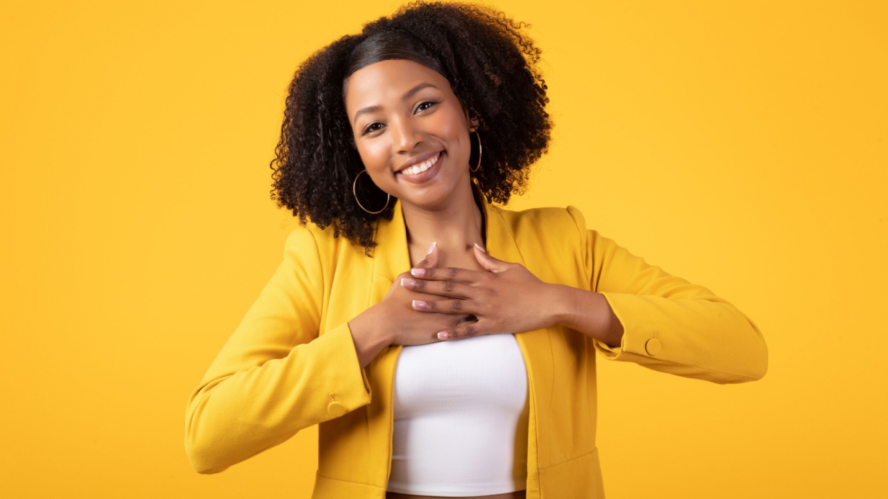 mulher negra com a mão no peito sorrindo vestindo amarelo em fundo amarelo