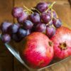 fotografia de um pote de vidro com duas romãs e uvas em cima de uma mesa de madeira