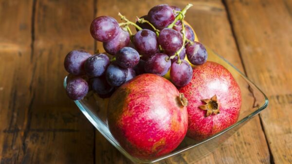 fotografia de um pote de vidro com duas romãs e uvas em cima de uma mesa de madeira