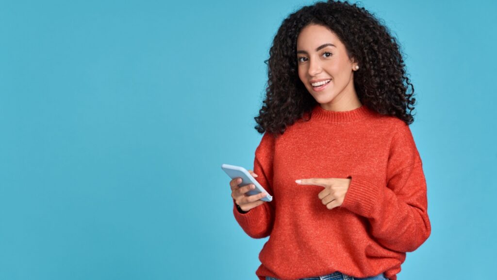 mulher segurando um celular na direita e apontando o dedo indicador da mão esquerda para ele em um fundo azul