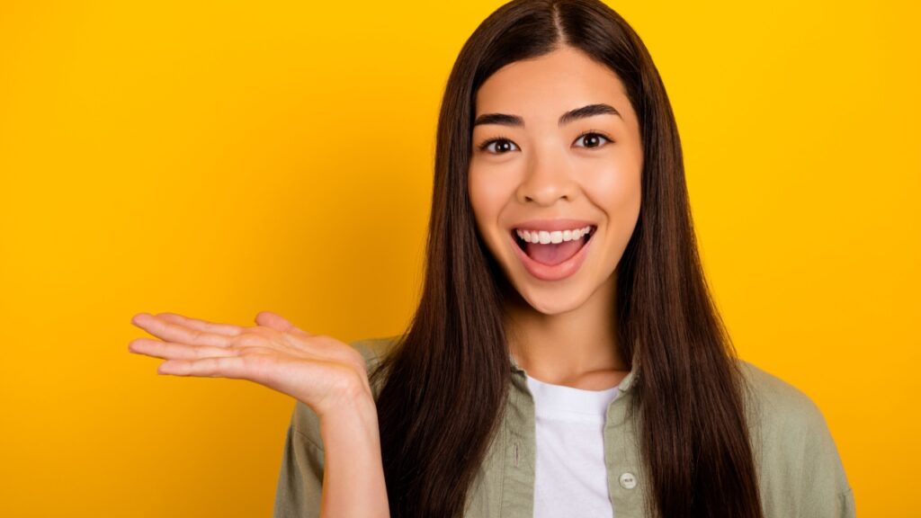 mulher com sorriso largo e a mão esticada na altura no pescoço em fundo amarelo