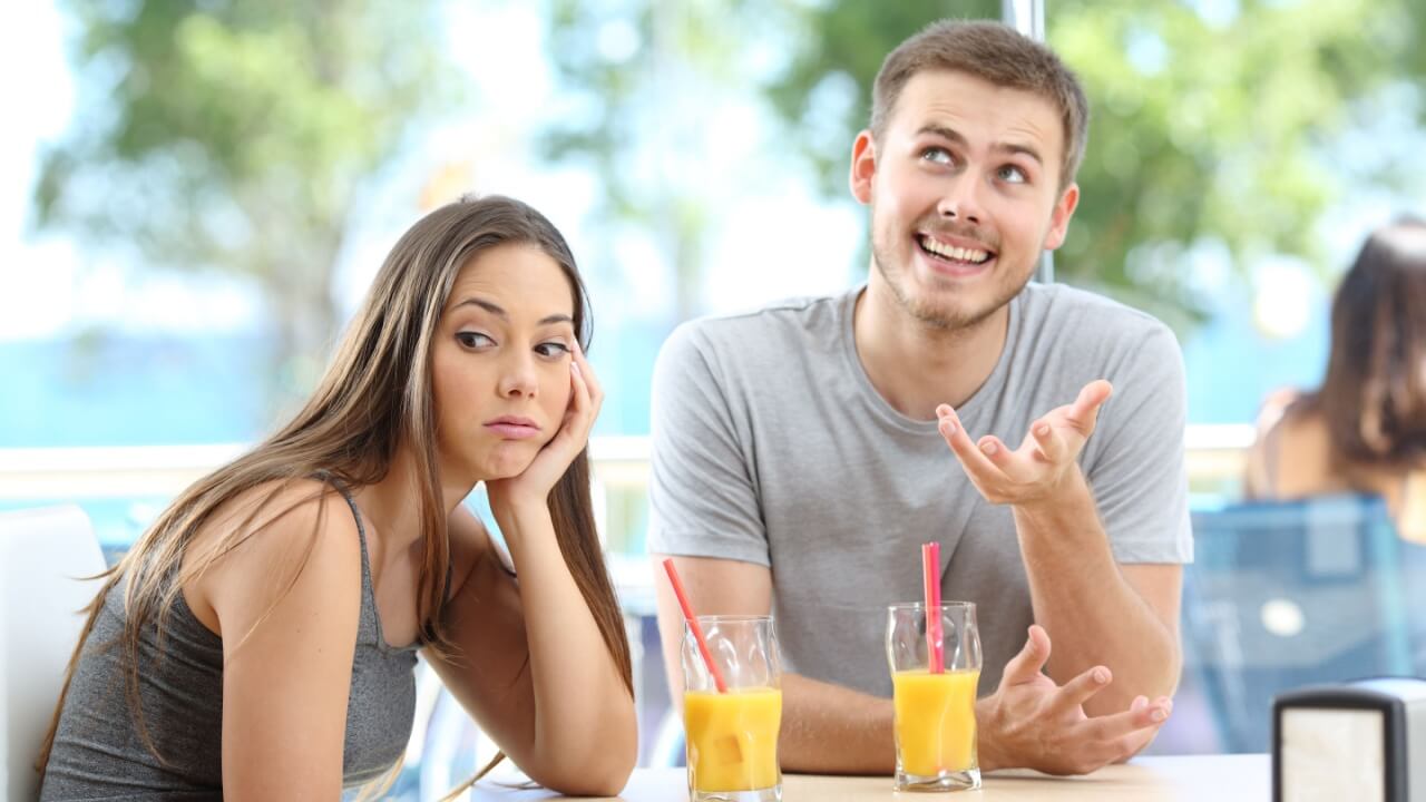 casal sentados em um restaurante com ele feliz e falando e ela com expressão de cansada