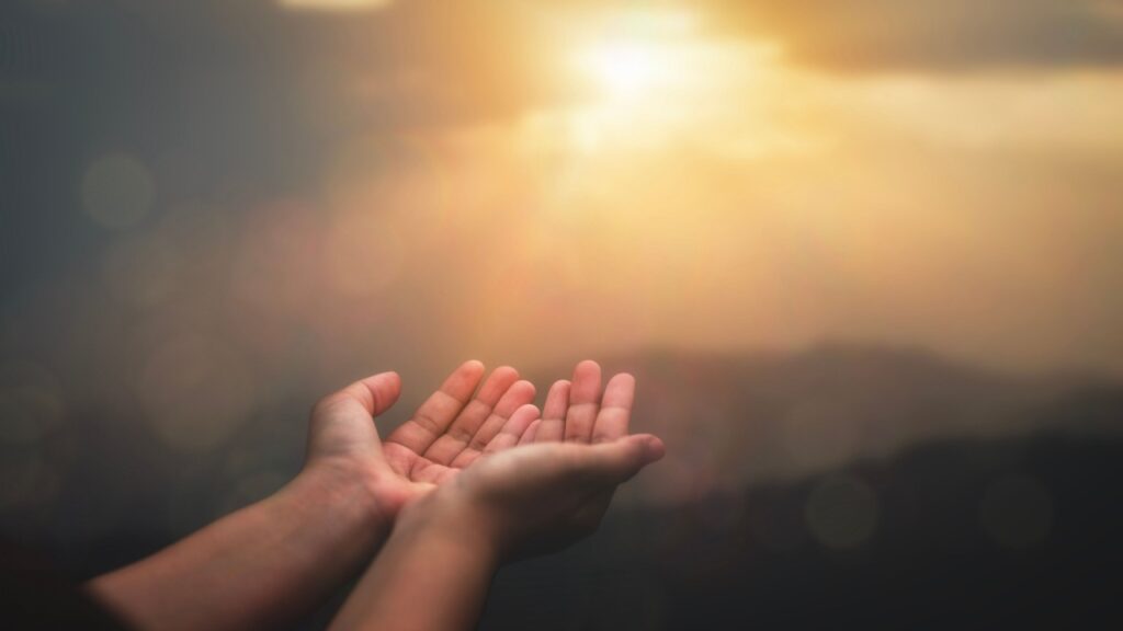 mãos esticadas em oração com raios de sol ao centro iluminando a imagem