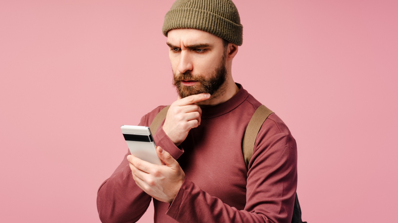 homem com uma mochila nas costas olhando para o celular com cara de dúvida em fundo rosa