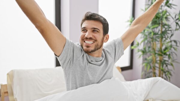 homem despertando na cama em um quarto bastante iluminado pela luz do sol e ele sorri largo